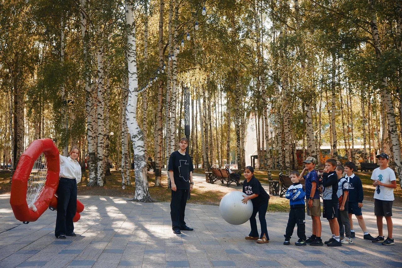 В лесопарке на дорожке стоят дети в синей форме. Перед ними женщина и мужчина. Слева стоит красный большой надувной предмет. Рядом надувной белый шар. Все смотрят на представление/игру. На деревьях осенняя листва.