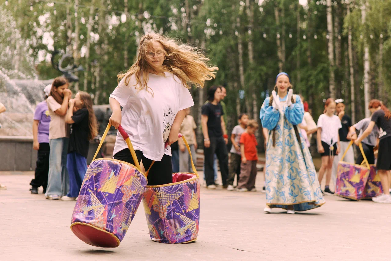 На улице в парке девочка в светлой футболке бежит и смеется.   У нее в руках две большие цветные сумки-ведра с широкими плетеными краями.   Сзади другие дети и взрослые стоят и тоже идут по дорожке, вокруг деревья и солнечный свет.
