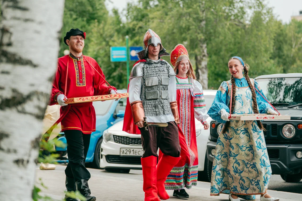 На фото группа людей на улице в национальных костюмах. Спереди человек в сером шлеме и жилете идёт по дороге, рядом идут женщины в ярких платьях и платках, за ними видна легковая машина и деревья вокруг.
