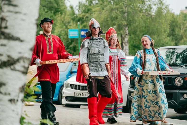 На фото группа людей на улице в национальных костюмах. Спереди человек в сером шлеме и жилете идёт по дороге, рядом идут женщины в ярких платьях и платках, за ними видна легковая машина и деревья вокруг.
